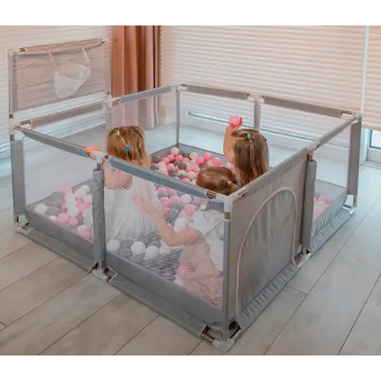 Lekegrind med ballbinge, grå nettingpaneler og rosa-hvite baller, tre barn leker inni, sett skrått ovenfra.