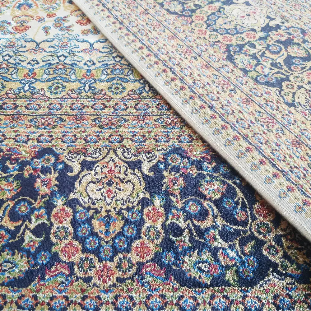 Qum Silk Tepe, detaljbilde av kremfarget teppe med blått og rødt orientalsk mønster, brettet hjørne viser veving.