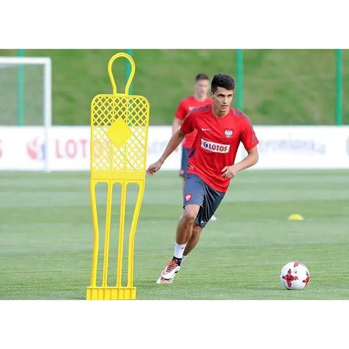 Gul treningsfigur i plast på gressbane, brukt under fotballtrening med spiller i rød drakt i bakgrunnen.