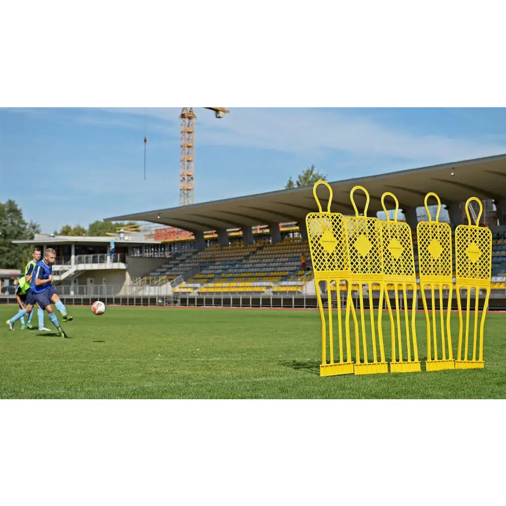 Fem gule treningsfigurer i plast på gressbane, sidevisning med fotballspillere og tribuner bak.