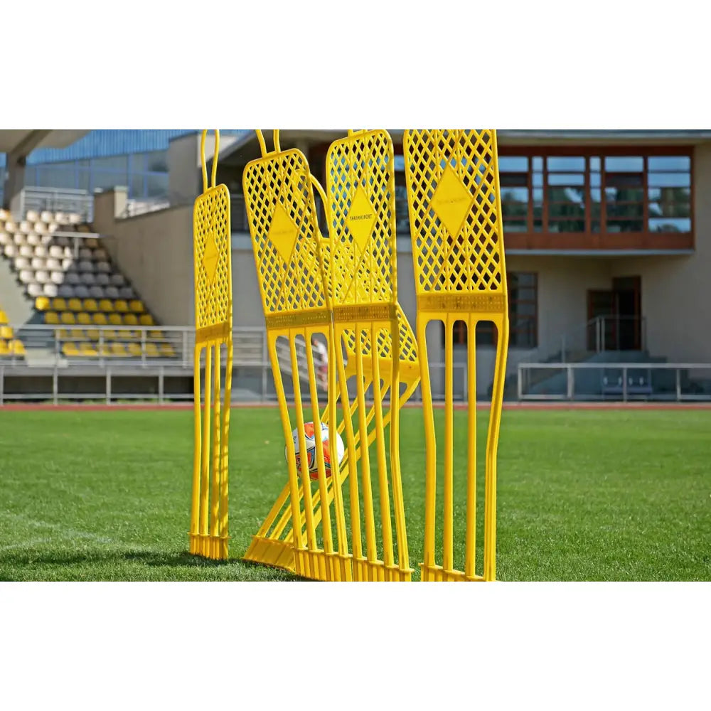 Mål Fodboldnett ELITE 180 cm Rød, fire gule plastmannekenger på rekke med fotball mellom, sett fra siden på gressbane.