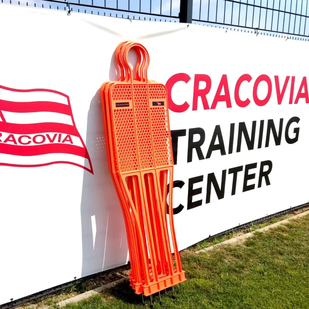 Målstolpe PRO 180 cm i oransje plast, fire stablede figurer foran gjerde på gress med skilt bak.