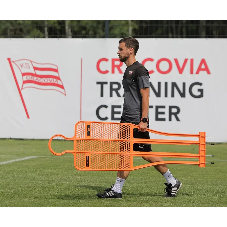 Målstolpe PRO 180 cm nylon, to oransje perforerte figurer bæres av mann på gressbane ved treningssenter.