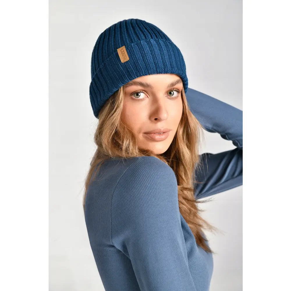 Blå merinoull lue med ribbestrikk og skinnlapp, nærbilde på kvinne fra siden mot lys bakgrunn.