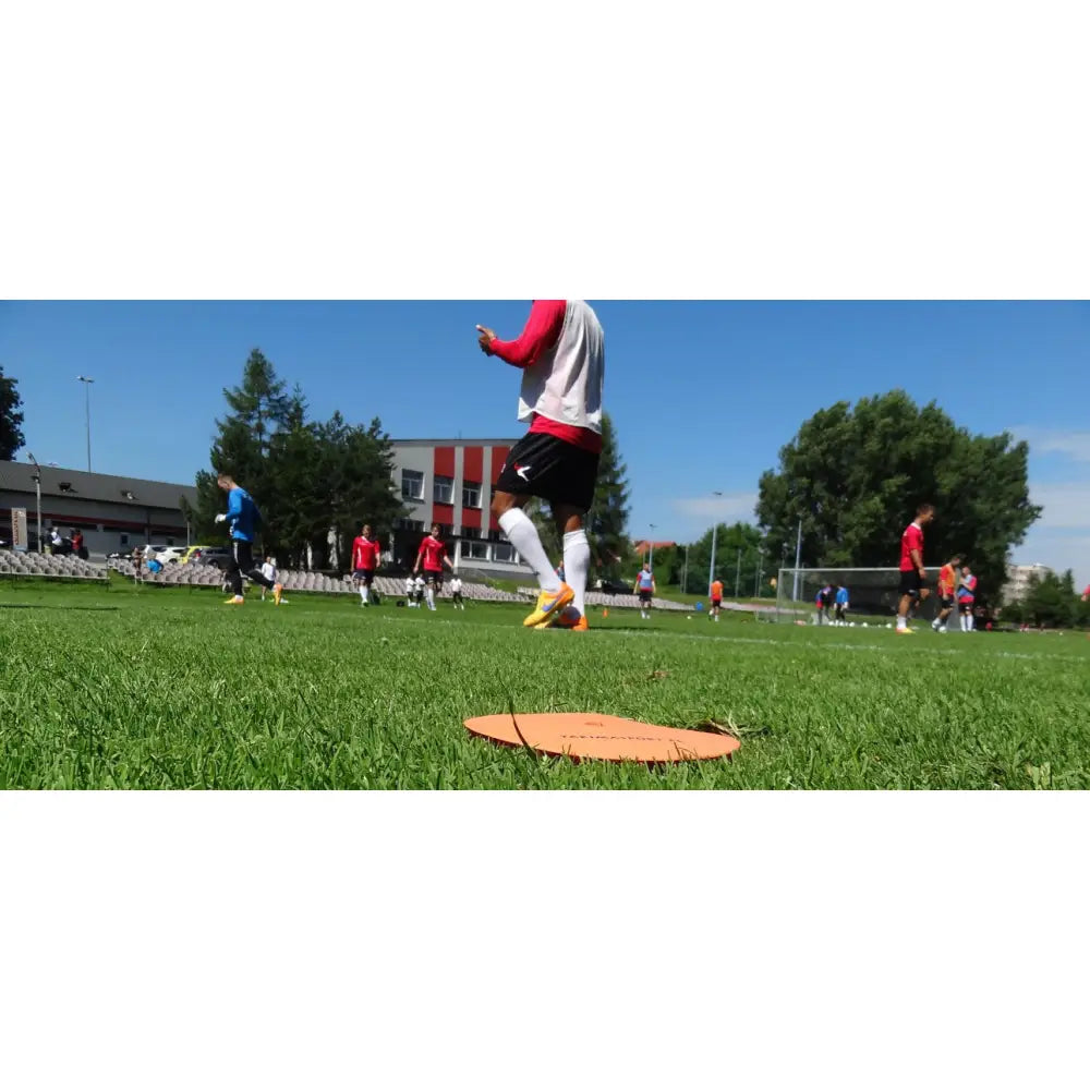 Oransje Merk Flat Marker markeringsbrikke på gress, nærbilde med fotballspillere i trening bak.