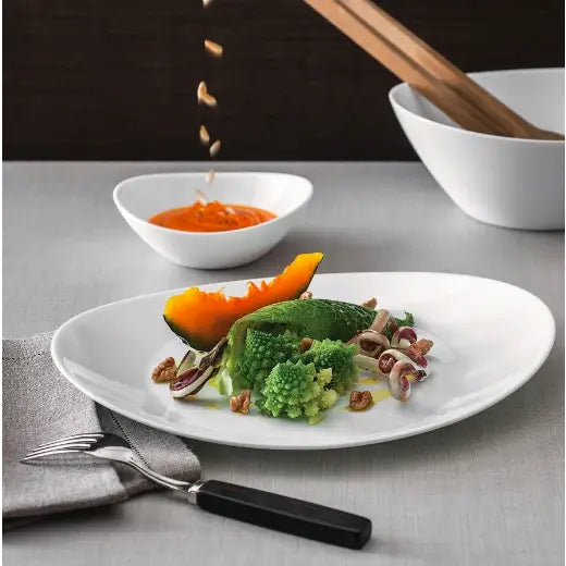 Oval hvit middagstallerken fra Prometeo, servert med grønnsaker og nøtter på grått bord, bestikk og serviett.