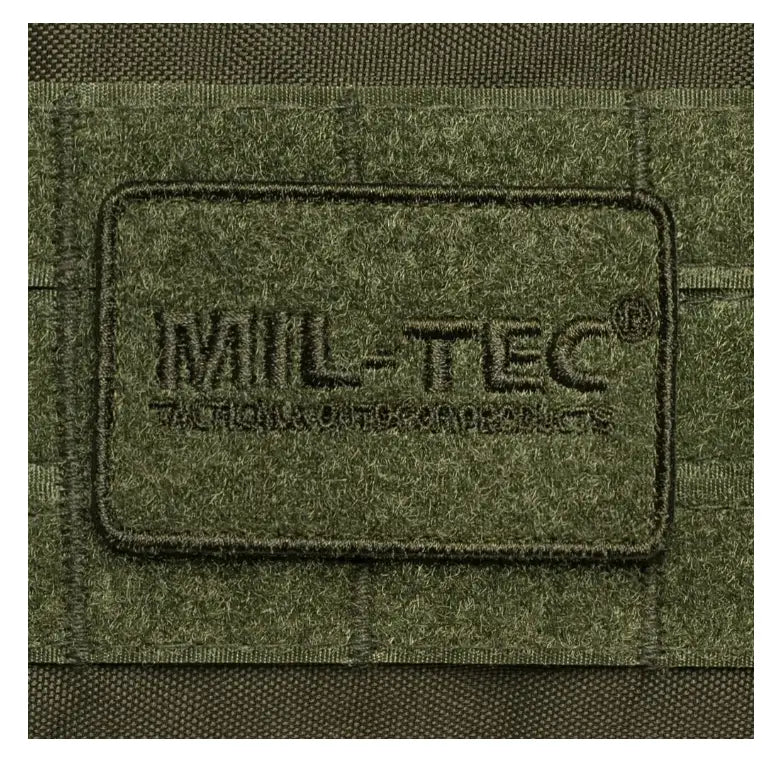 Olivengrønn borrelåslapp med sort Mil-Tec-logo, detaljvisning av tekstil og søm på ryggsekk.