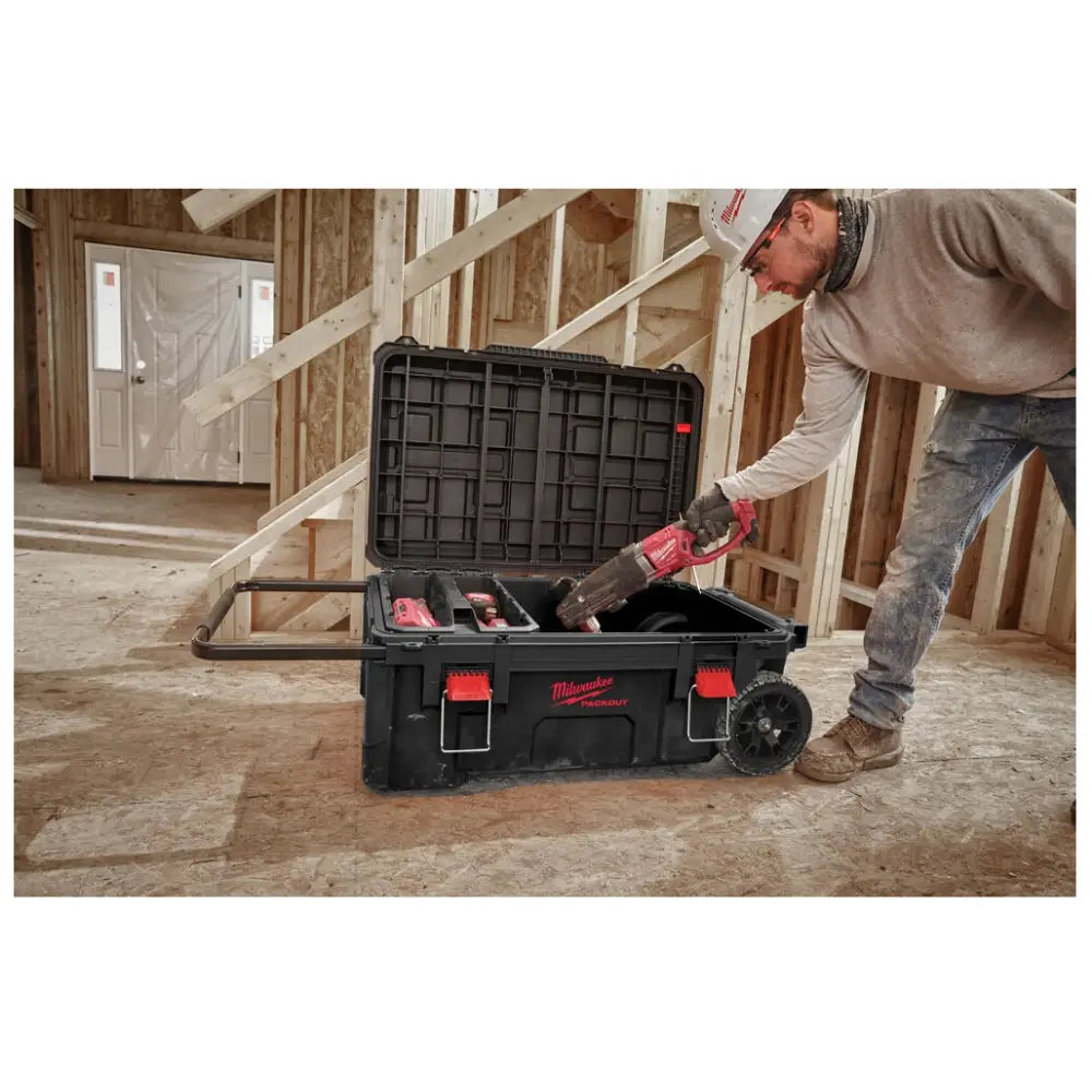 Milwaukee Packout kasse på hjul, åpen med rød drill plassert oppi av en person på byggeplassgulv.