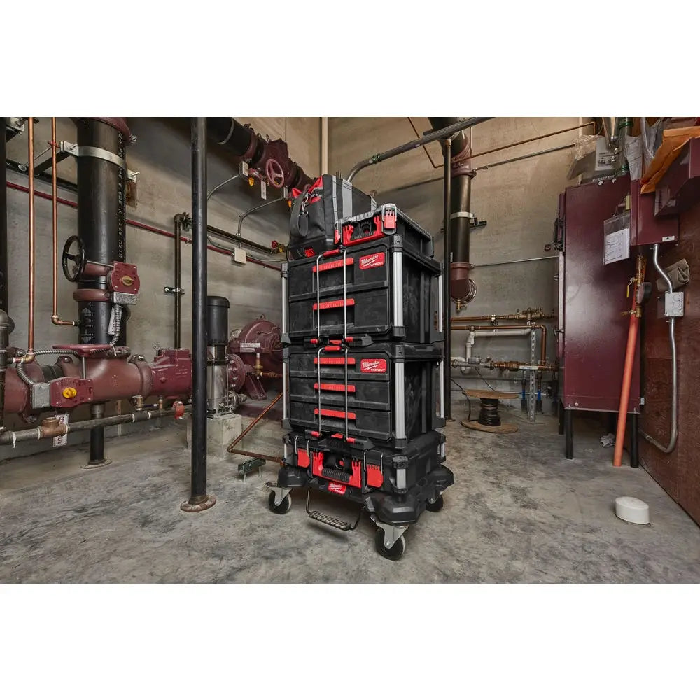 Milwaukee Packout verktøykasse, fire kasser stablet på tralle med hjul i fabrikkmiljø med rør og maskiner.