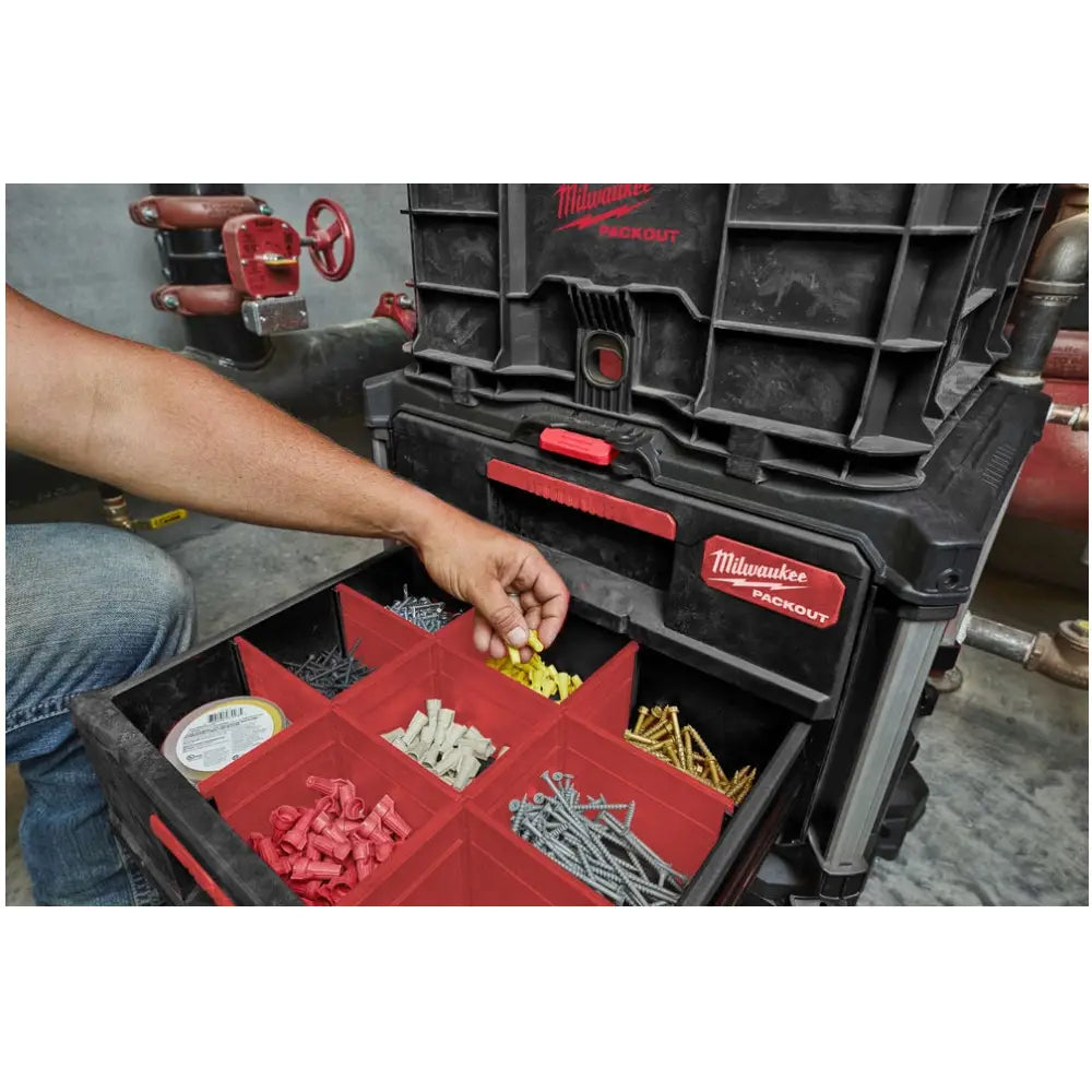 Milwaukee Packout verktøykasse åpen, hånd tar ut gule plugger fra rødt rom blant sorterte skruer og deler.