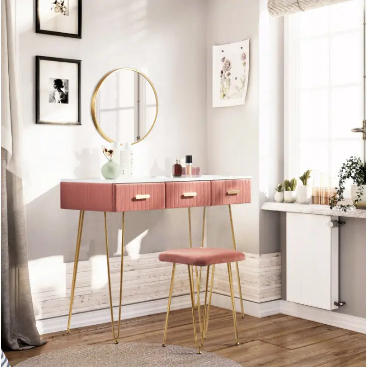 Sminkebord med tre rosa skuffer, gullben og rundt speil, vist med matchende rosa krakk i lyst soverom.