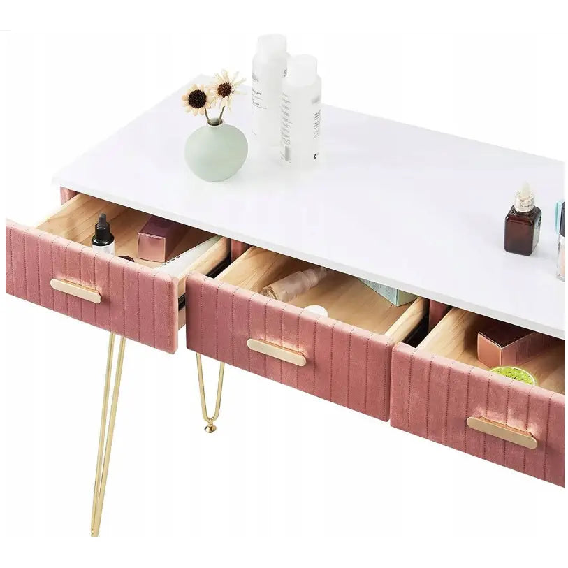 Moderne sminkebord med hvit plate, tre rosa fløyelsskuffer og gulldetaljer, sett skrått med sminkeprodukter.