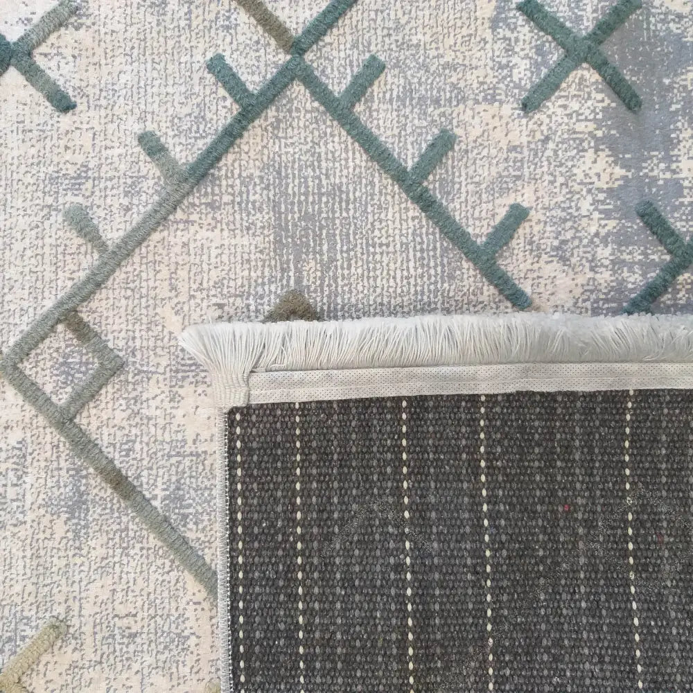 Moderne teppe Hypnotisk 02, nærbilde av hjørne med blågrønne linjer, grå bakside og hvite frynser.