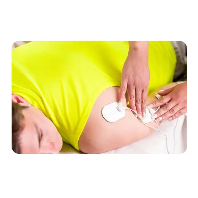 Muskelstimulator med to elektroder plassert på skulderen til person i gul t-skjorte, ledninger synlige.