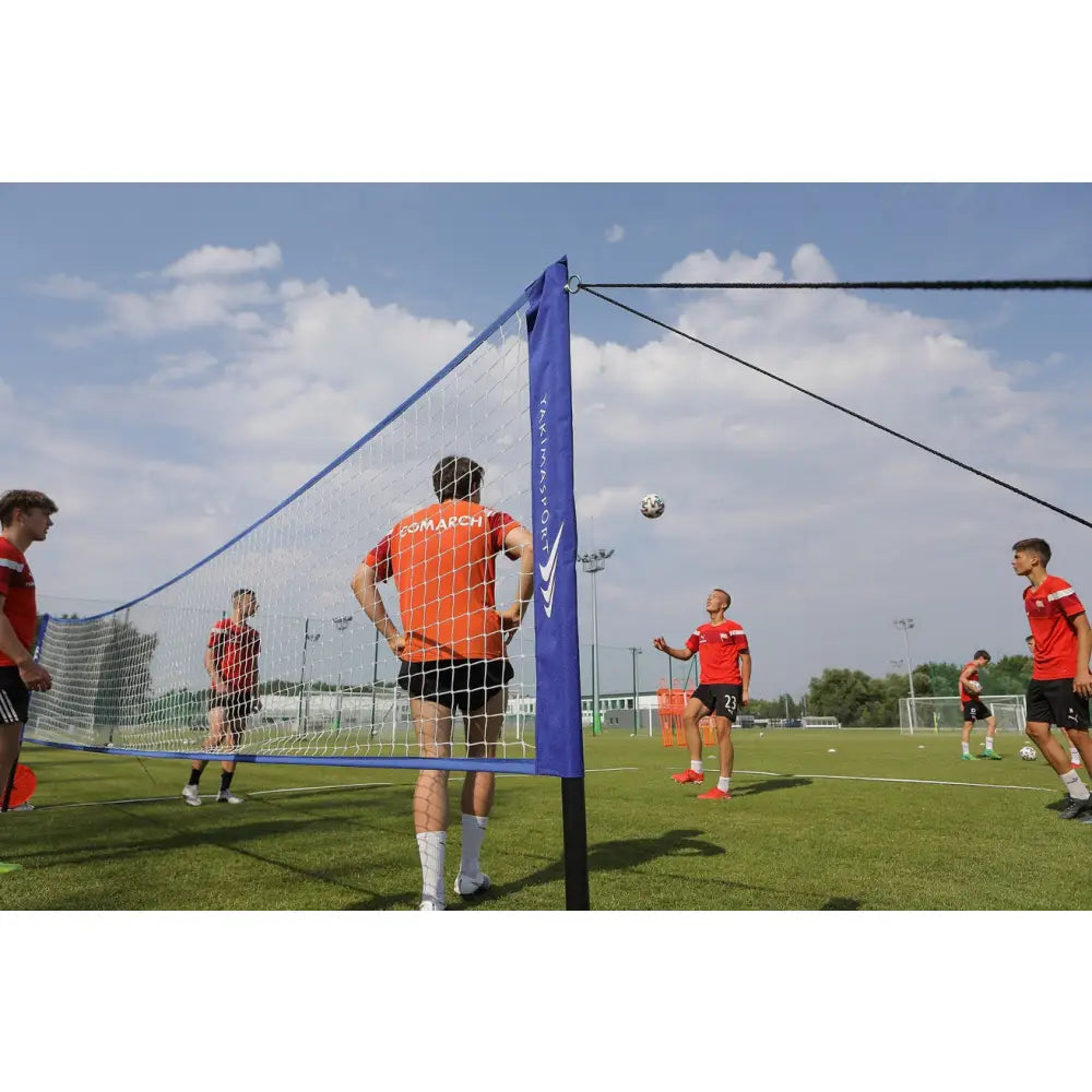 Sportnett med blå kant og sort stolpe, sidevisning på gressbane under fotballtrening med rødkledde spillere.