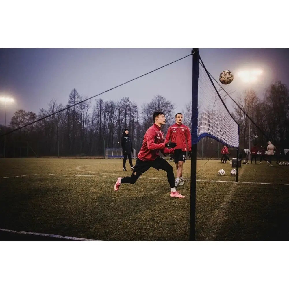 Naturlig gressmatte, to unge menn i røde jakker spiller fotball over nett på flombelyst gressbane i skumringen.