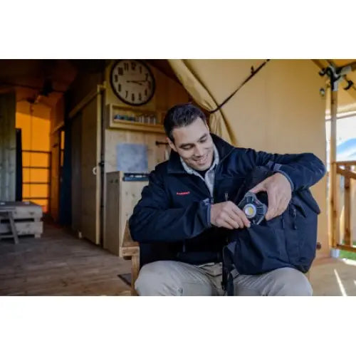 Oppladbar arbeidslampe LED, sort og gul modell løftes ut av veske av mann foran veggklokke og treinteriør.