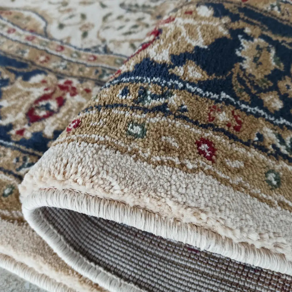 Ovalt kremfarget teppe med marineblå, gull og røde detaljer, sammenrullet for å vise luv og kantfinish.