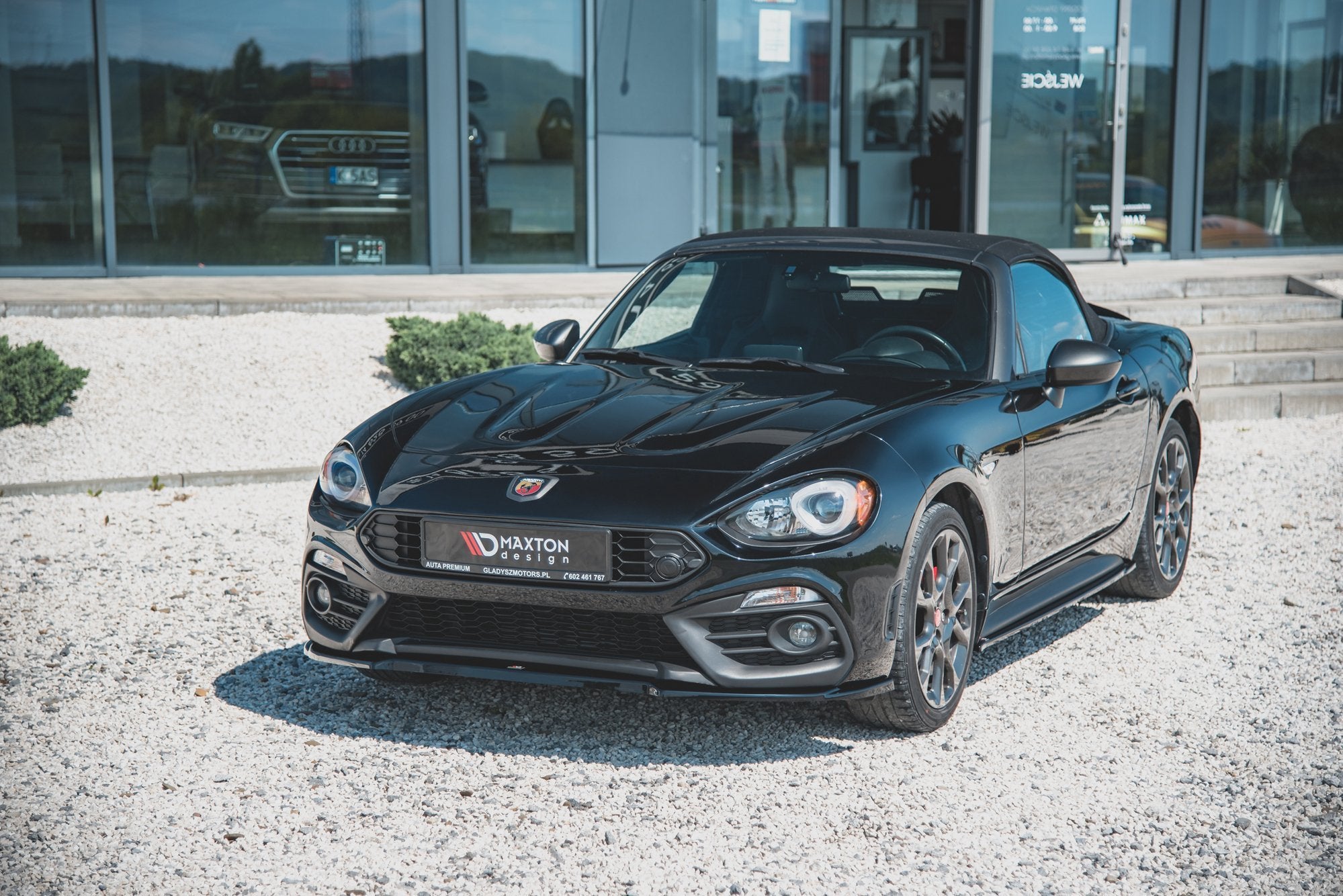 Frontspoiler på svart Abarth 124 Spider med Maxton Design-logo, parkert på grus foran bygning.
