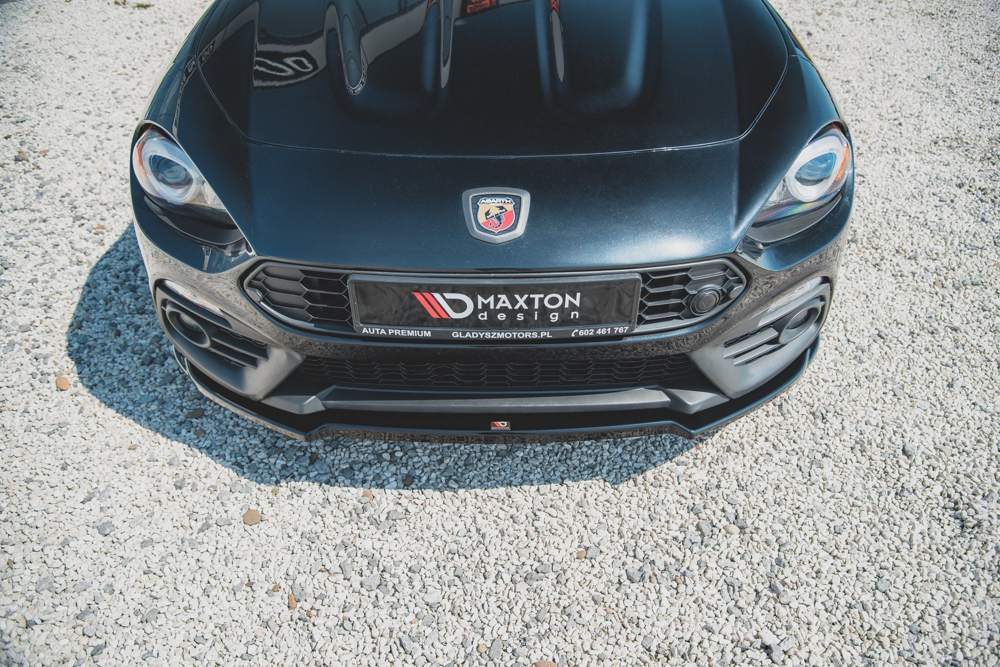 Frontleppe på Abarth 124 Spider med Maxton Design-skilt, fotografert skrått ovenfra på grus.