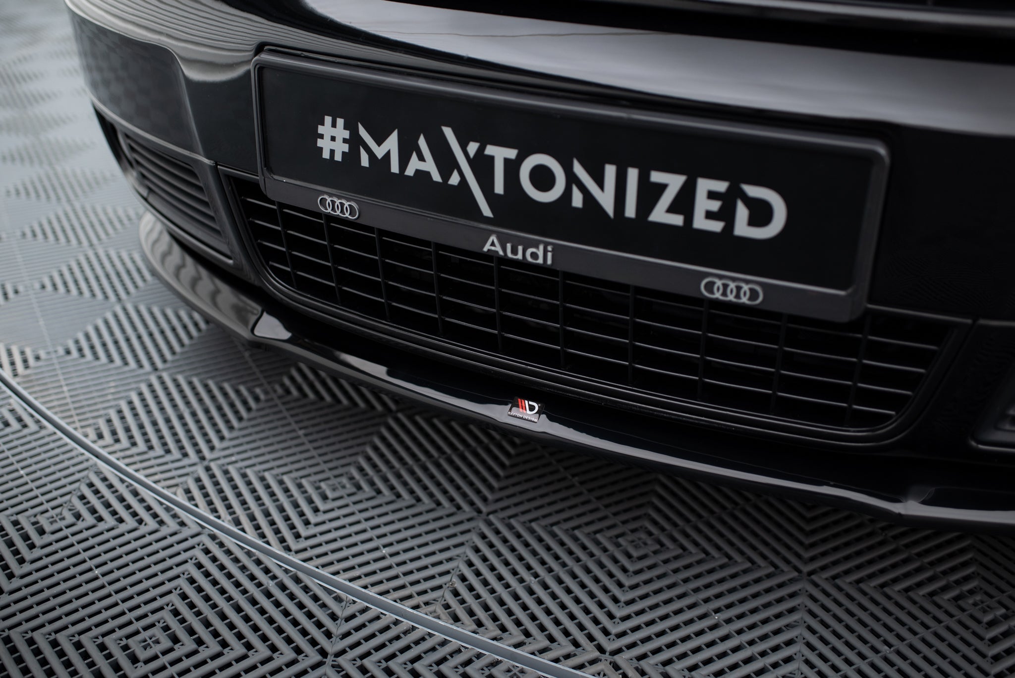 Spoiler til Audi S6 C5 V2, sort blank plast sett forfra med logo og bilens grill synlig over.