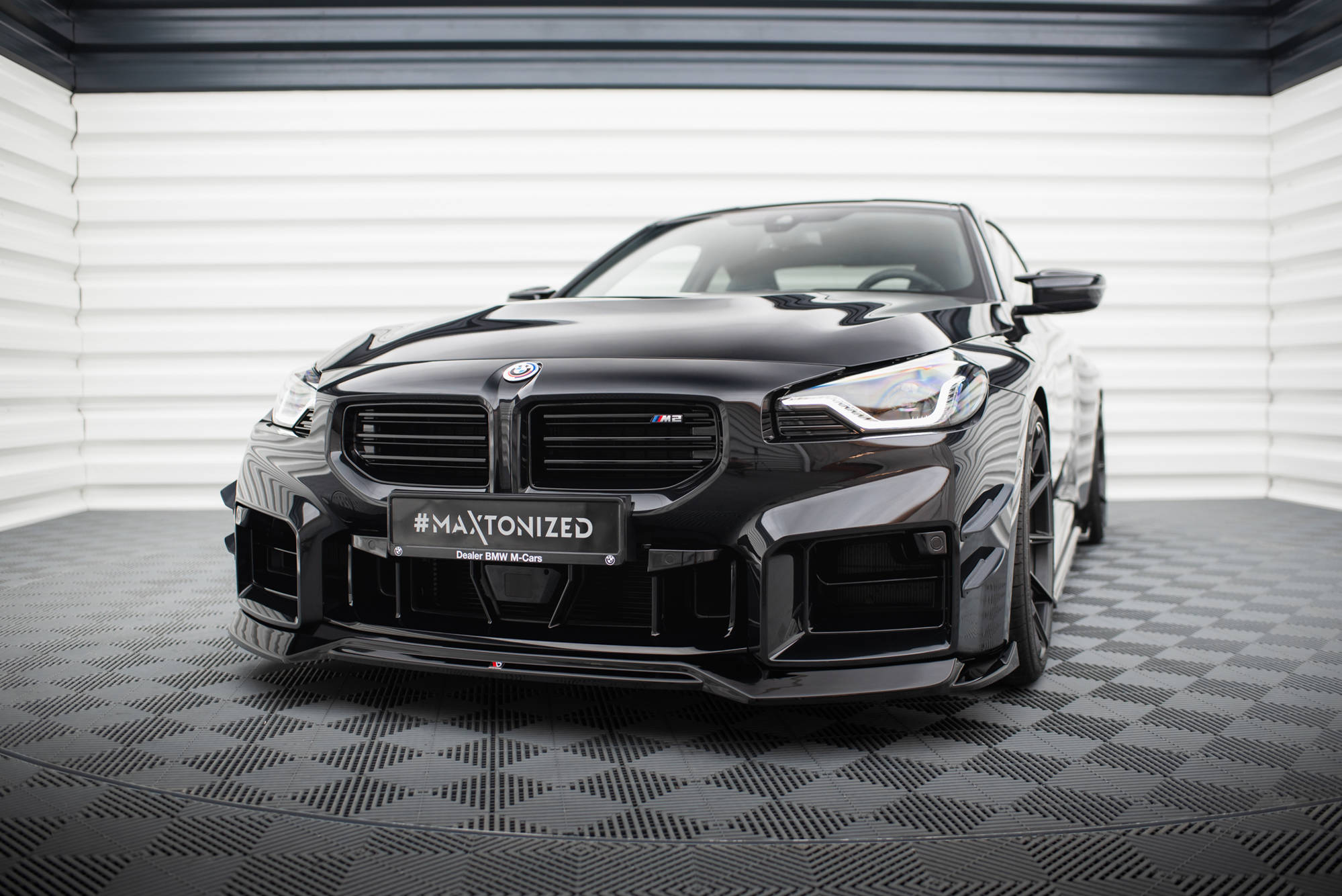 Frontspoiler BMW M2 G87 V.1 montert på sort sportsbil med brede felger og LED-frontlys, innendørs.