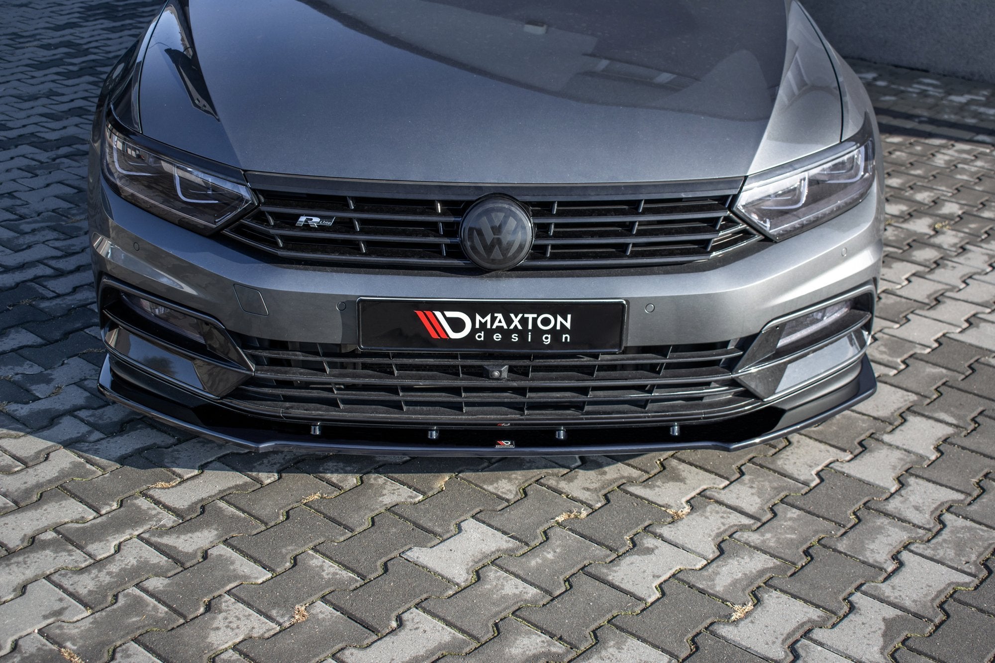 Frontspoiler VW Passat R-Line B8 V.1 montert på grå bil med sort grill og mørke hovedlykter, sett forfra.