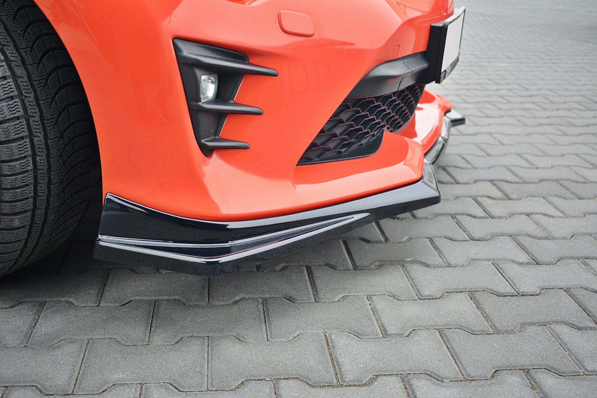Front splitter på oransje Toyota GT86 Facelift TUV V.2 med matt svart plastdetaljer på grå belegningsstein.