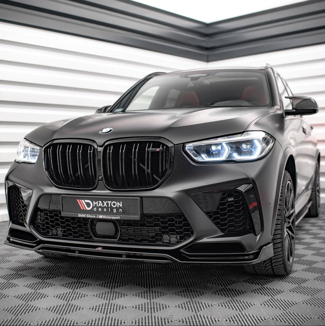 Frontspoiler BMW X5M F95 V.3 montert på sort SUV med sportslig frontfanger og LED-lys i lyst rom.