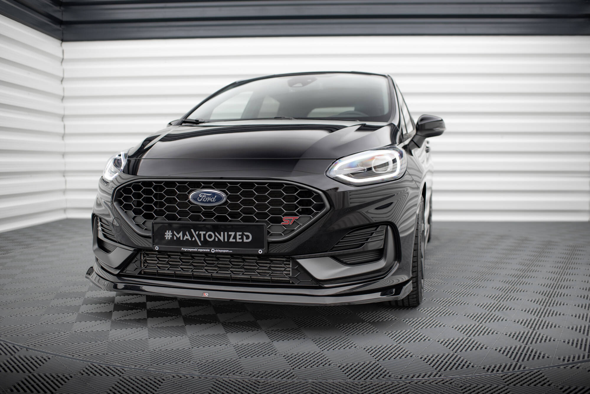Frontspoiler til Ford Fiesta MK8 Facelift ST med svart grill og LED-lykter sett forfra innendørs.