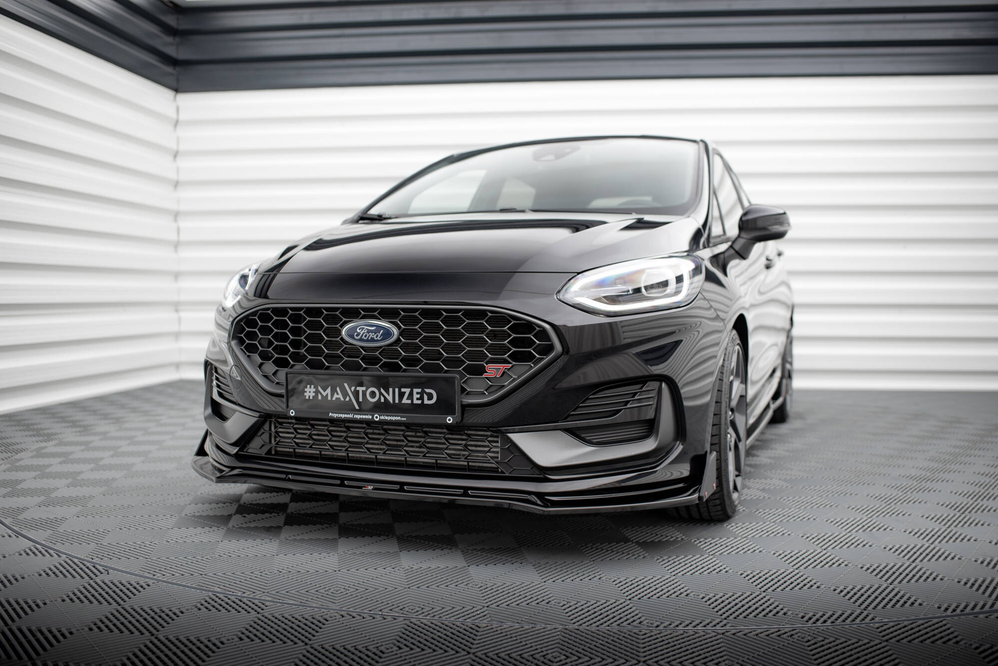 Frontspoiler Ford Fiesta MK8 Facelift St vises montert på svart bil med sportsgrill i lyst industrilokale.
