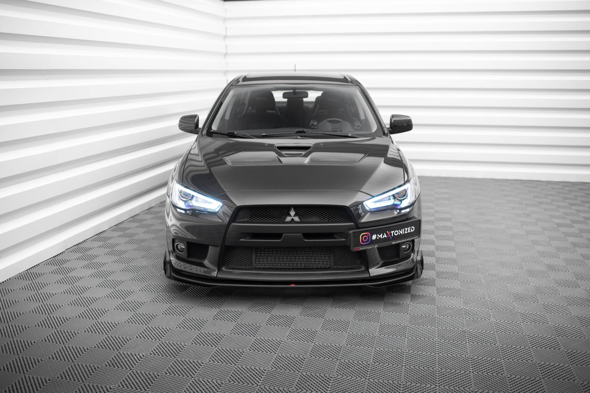 Frontspoiler Mitsubishi Lancer Evo X med flaps V1, sort sportsbil sett rett forfra i lyst rom.