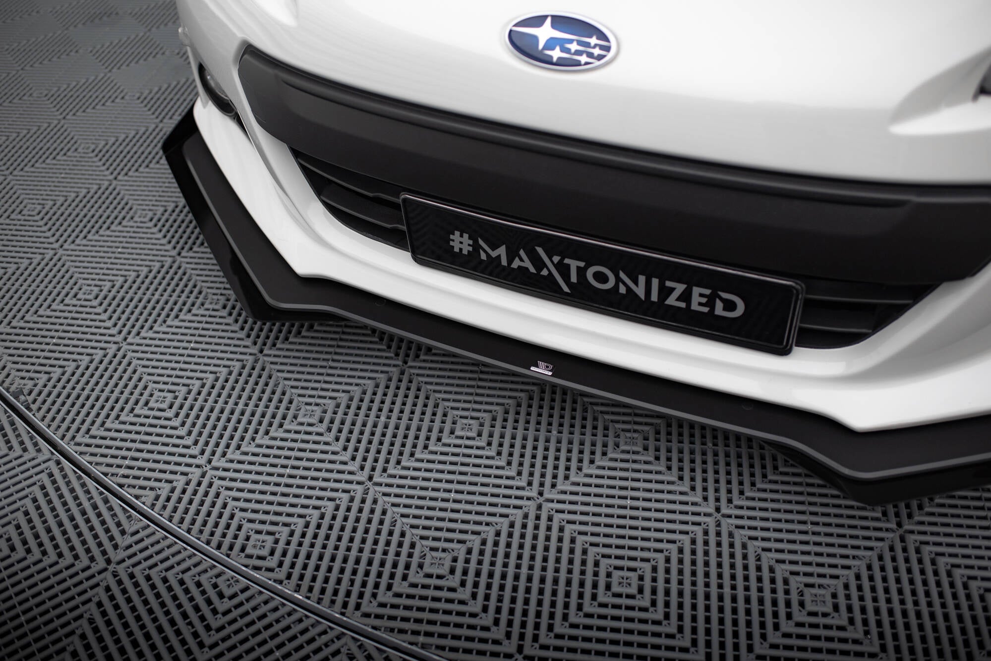 Spoiler til Subaru BRZ MK1 2012-2017 V2 V.1 i sort materiale, nærbilde sett forfra mot grått mønster.