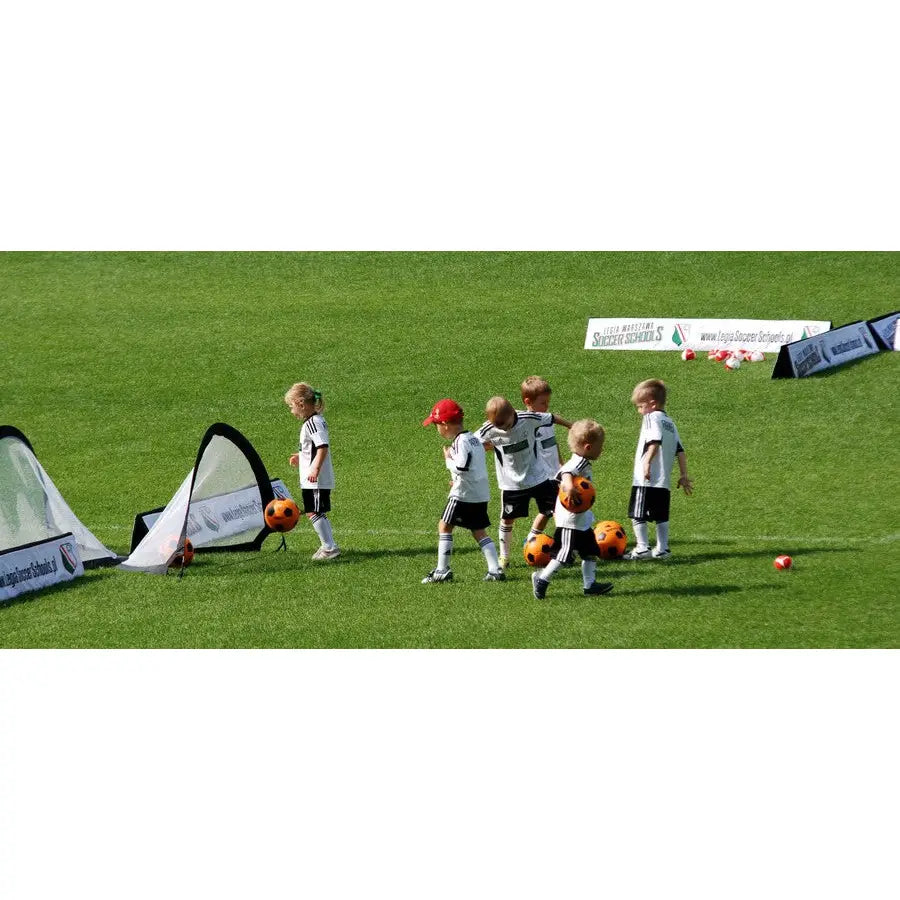 Pop-up portabelt fotballmål 120x80 cm, barn spiller fotball med oransje baller på gress, sideperspektiv.