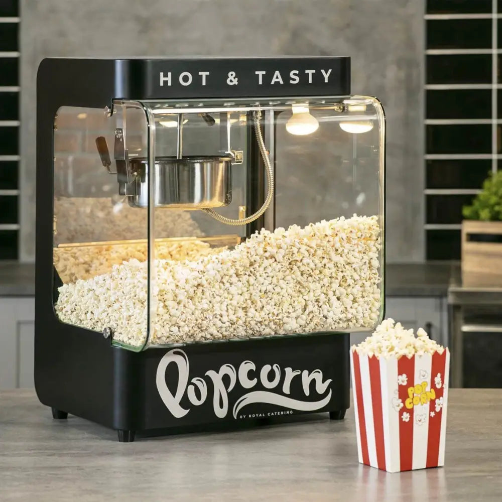 Svart popcornmaskin med glassvegger, synlig fersk popcorn, "HOT & TASTY"-tekst og popcornbeger ved siden av.