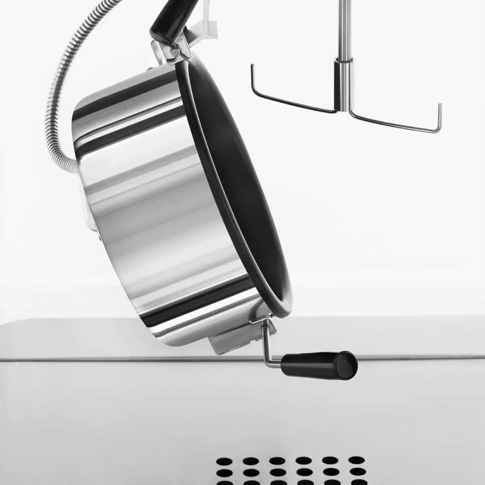 Poppemaskin i stål, nærbilde av sidevis kjel med sort håndtak og synlig vispetilbehør i bakgrunnen.