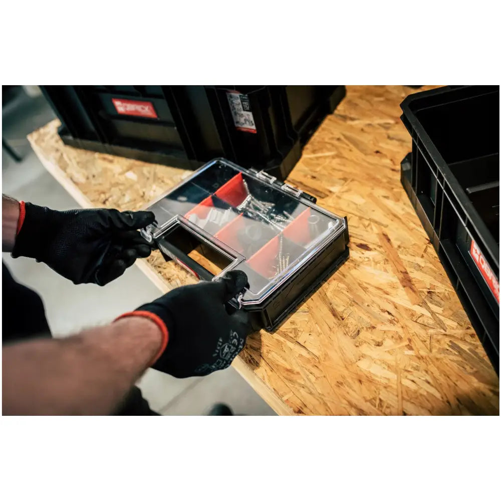 Qbrick verktøykasse sett med røde rom og gjennomsiktig lokk, åpnes av hender med hansker på arbeidsbord.