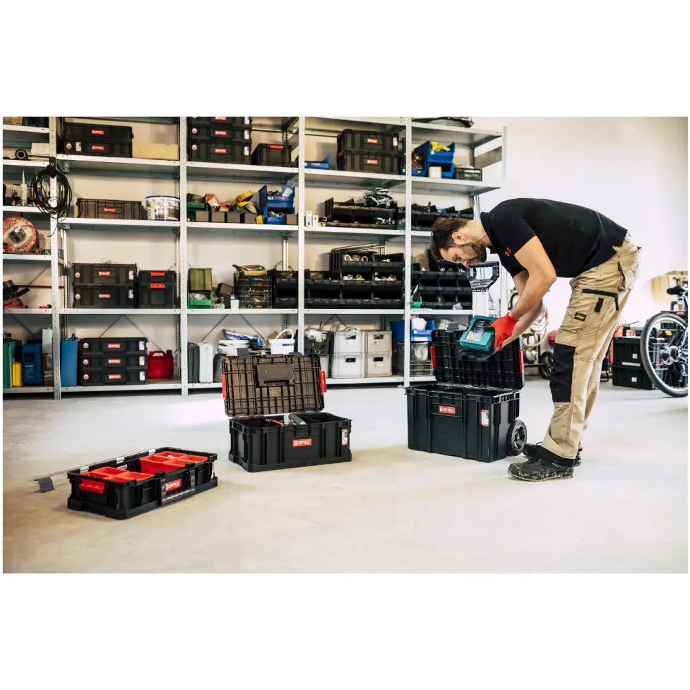 Qbrick verktøykasse sett Two, en mann sorterer verktøy i svarte kasser med røde detaljer foran hyller.