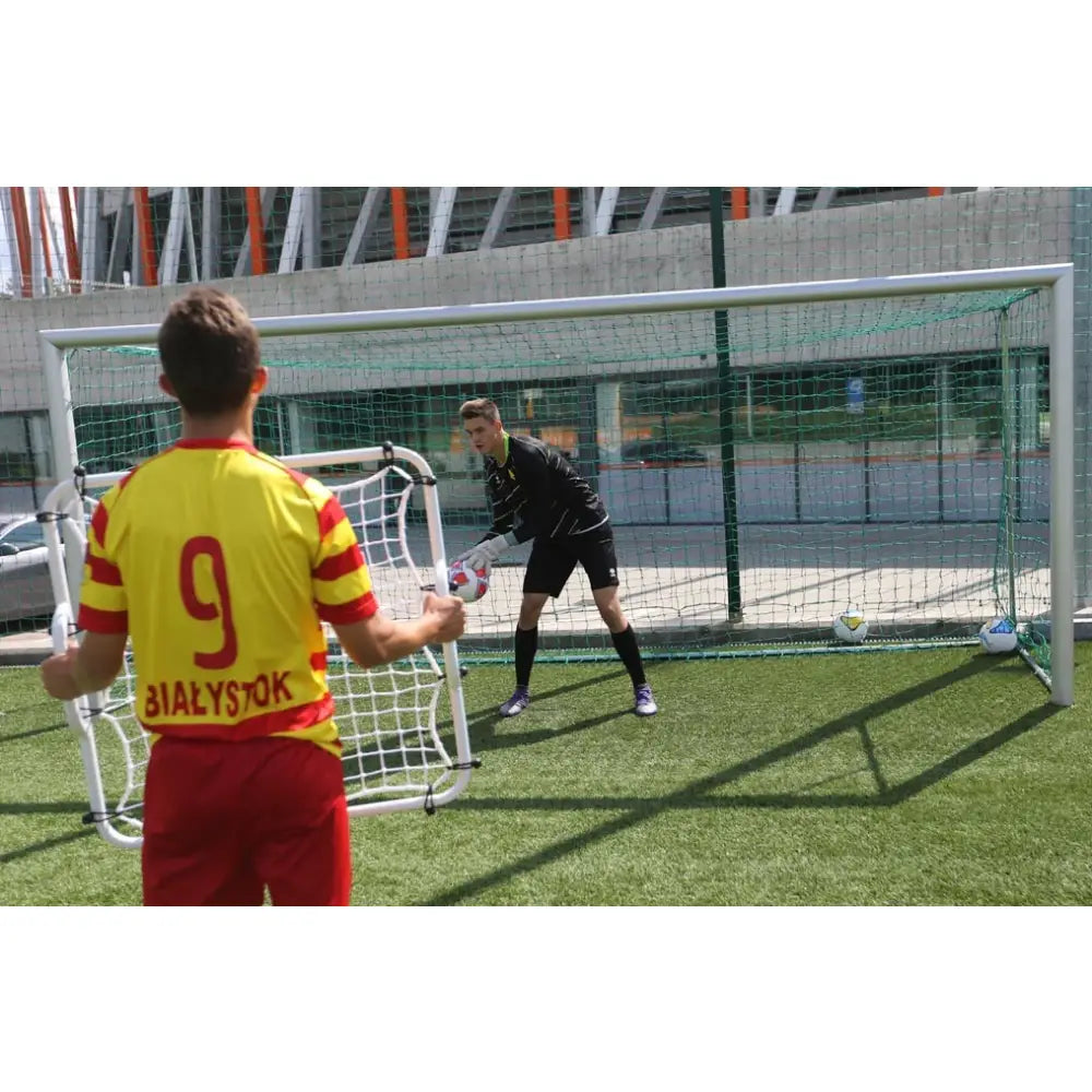 Rebounder MINI for keepertrening, brukt av spiller på kunstgress foran mål med keeper klar i bakgrunnen.