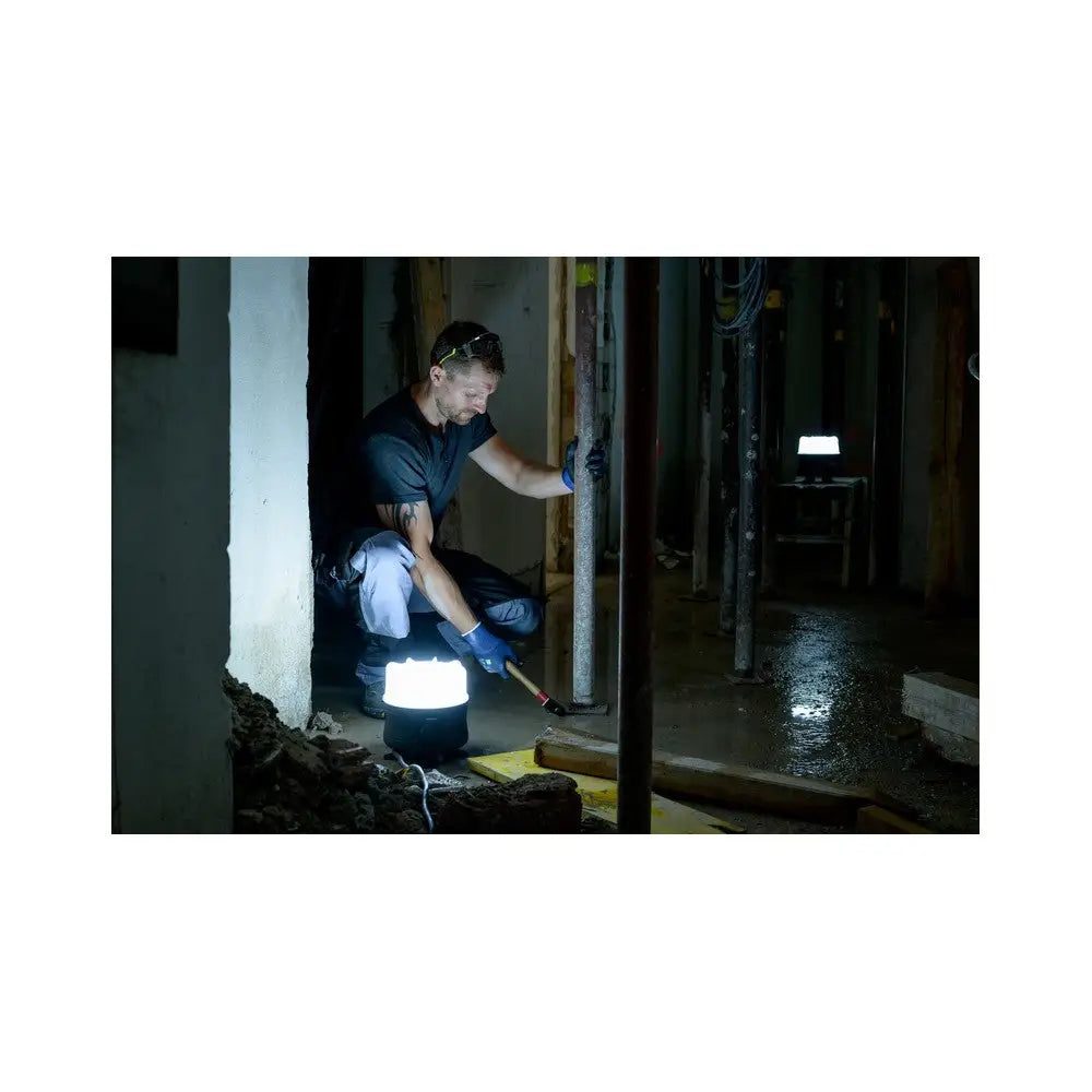 Reflektor LED 360°, arbeidslykt på gulv som lyser opp arbeider med brekkjern i mørkt byggmiljø.