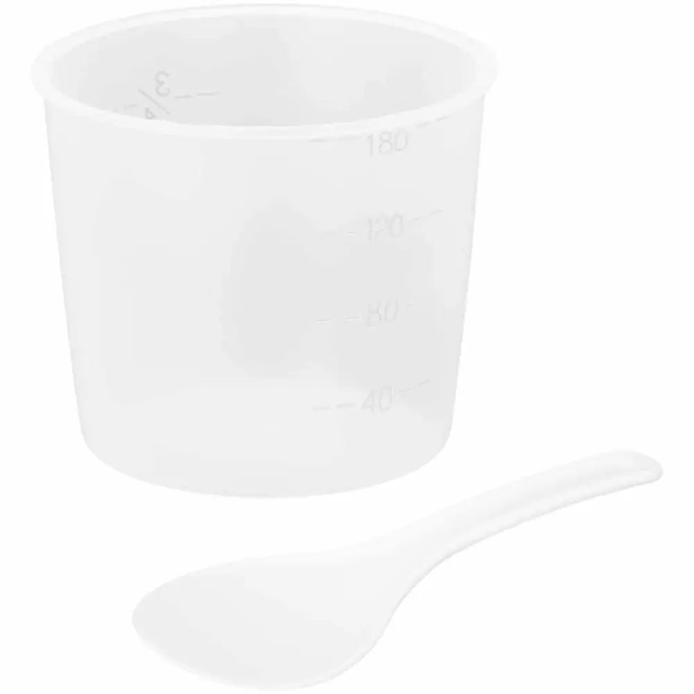 Transparent målebeger med millilitermarkeringer og hvit plastskje foran, sett fra siden mot hvit bakgrunn.