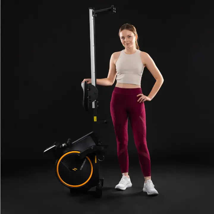 Romaskin Omega, sammenfoldet for oppbevaring, med kvinne i treningstøy ved siden av mot mørk bakgrunn.