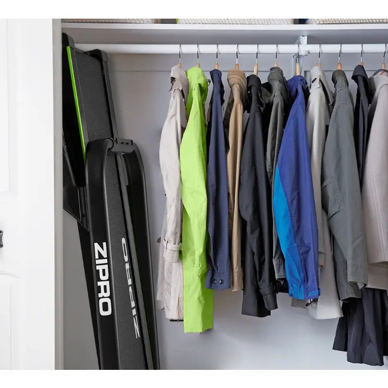 Sammenleggbar Tredemølle Zipro Jogger, sort modell oppbevart vertikalt i garderobeskap ved siden av fargerike jakker.