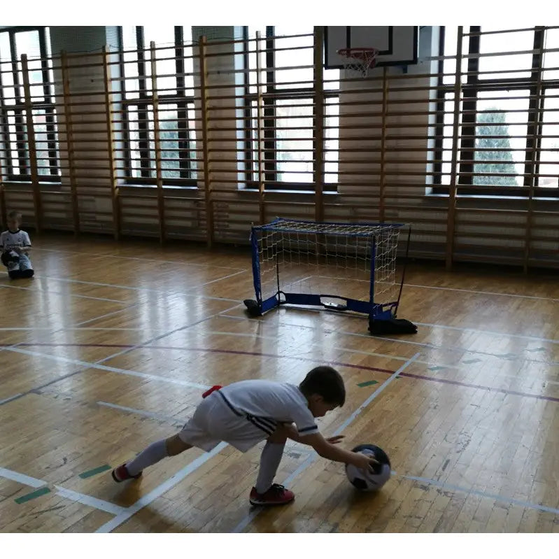 Sammenleggbart blått fotballmål 120x80 cm i gymsal, barn i hvit drakt skyter ball mot målet.