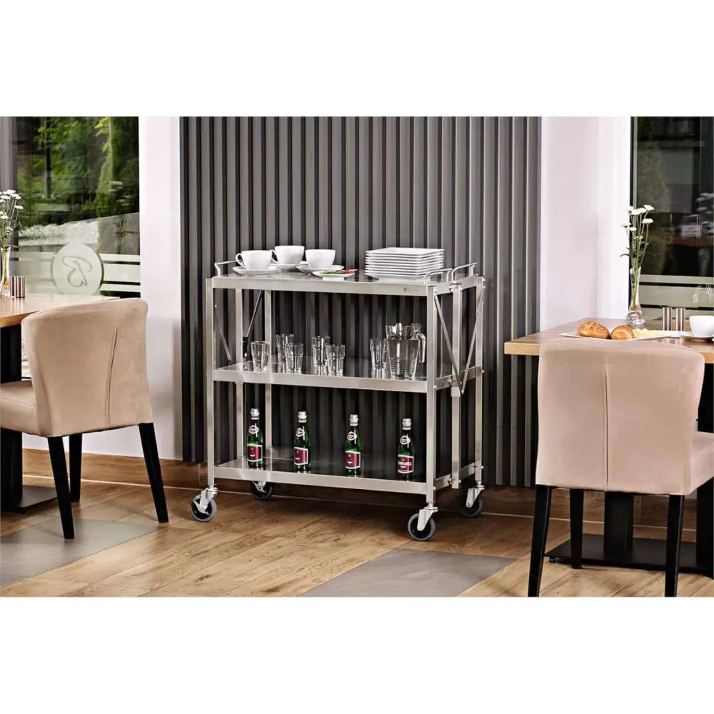 Sammenleggbar serveringsvogn i rustfritt stål med tre hyller, fylt med servise og flasker i restaurantmiljø.