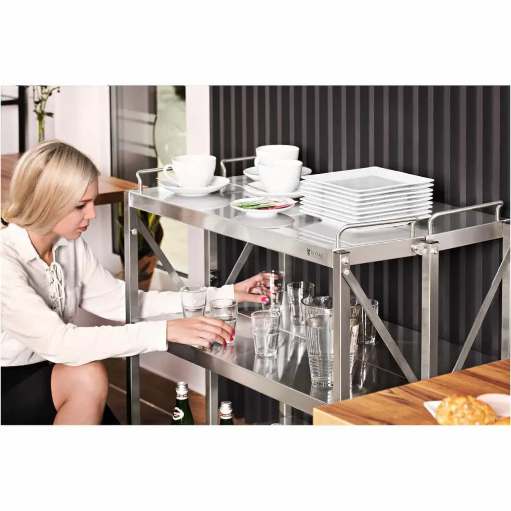 Serveringsvogn i rustfritt stål med tre hyller, sett fra siden mens glass plasseres på nederste hylle.