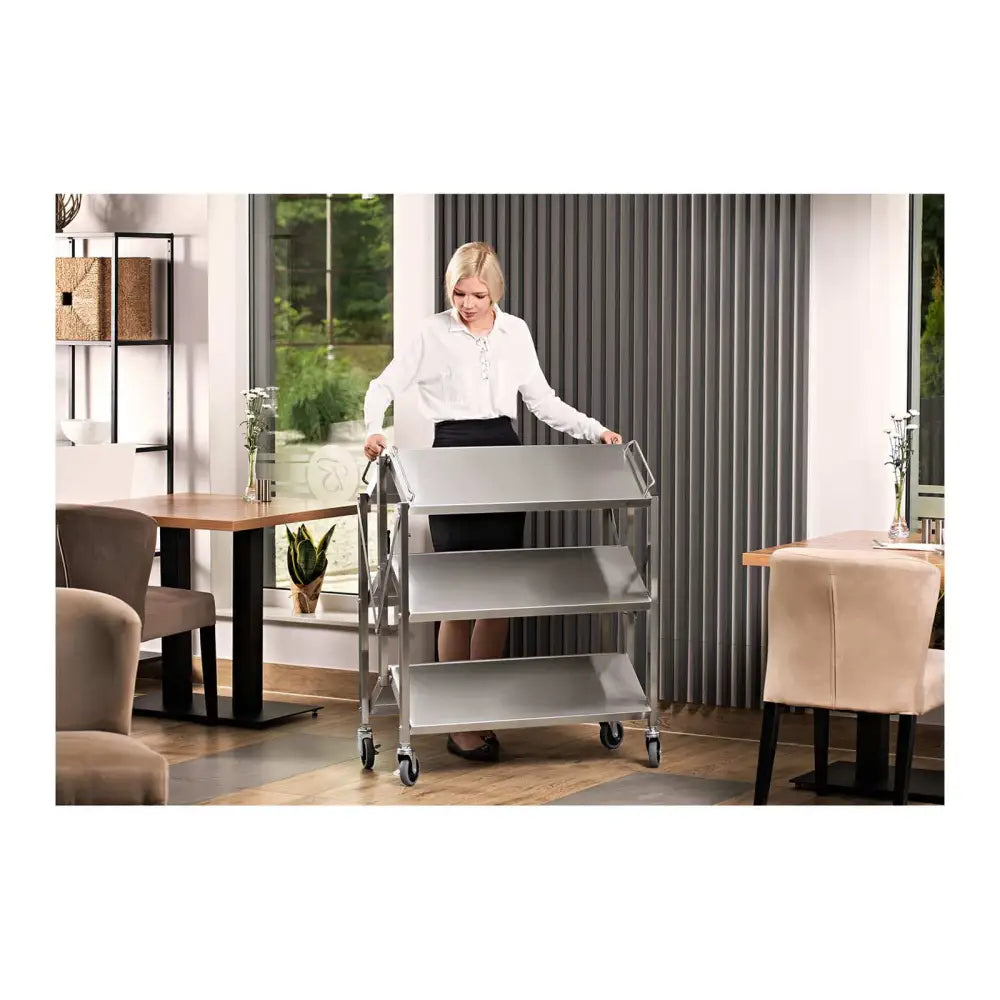 Serveringsvogn i rustfritt stål med tre hyller, vist i bruk av kvinne i restaurantmiljø, front/side.