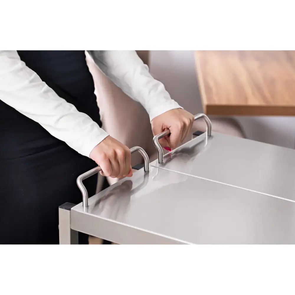 Serveringsvogn i stål med tre hyller, nærbilde av hender som holder i håndtakene under bruk.