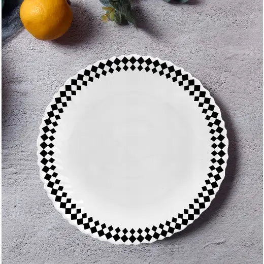 Hvit middagstallerken med bølget kant og svart-hvitt rutemønster, sett ovenfra på grå overflate.