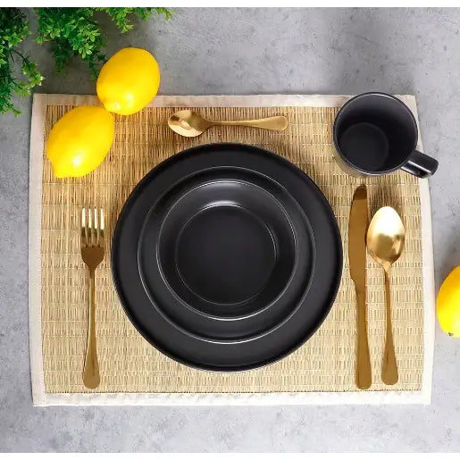 Servise i svart keramikk sett for fire, sett ovenfra med gullbestikk og sitroner på lys bordmatte.
